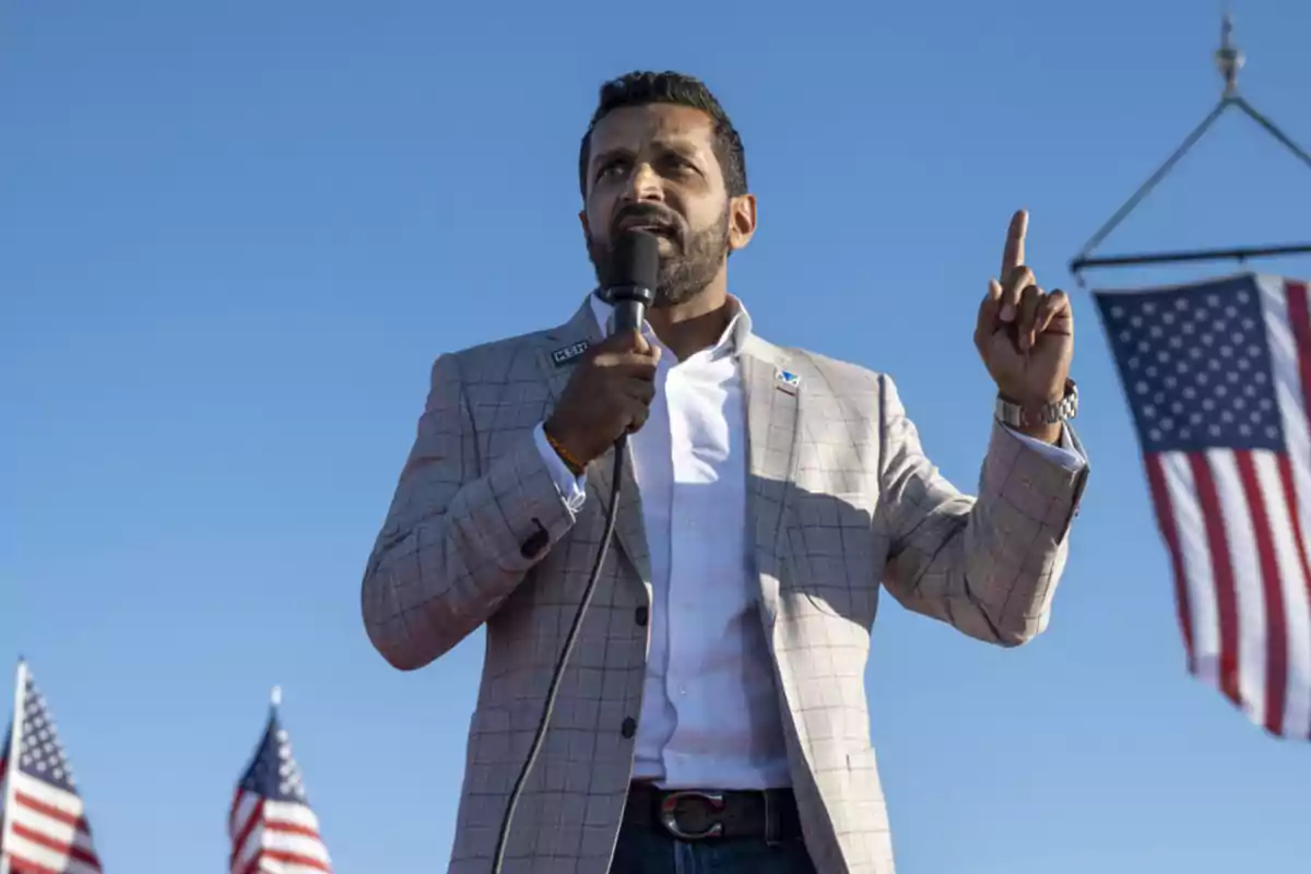 Hombre con barba hablando por micrófono y levantando un dedo durante un evento al aire libre con banderas de Estados Unidos al fondo Hombre con barba hablando por micrófono y levantando un dedo durante un evento al aire libre con banderas de Estados Unidos al fondo