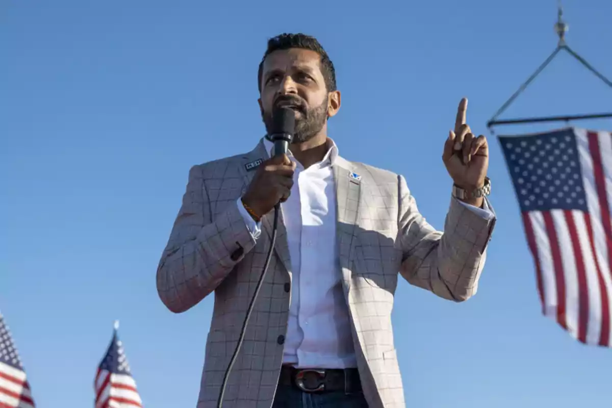 Man in a light-colored jacket speaking into a microphone outdoors with U.S. flags in the background Man in a light-colored jacket speaking into a microphone outdoors with U.S. flags in the background