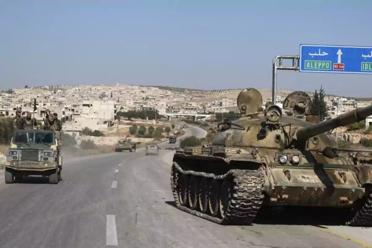 Un tanque y un vehículo militar circulan por una carretera en una zona urbana con un cartel que indica la dirección hacia Aleppo e Idlib.