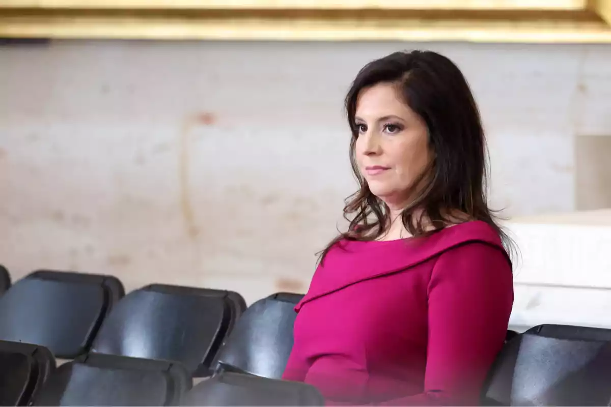 A woman in a fuchsia dress is sitting in a row of black chairs in an indoor setting. A woman in a fuchsia dress is sitting in a row of black chairs in an indoor setting.