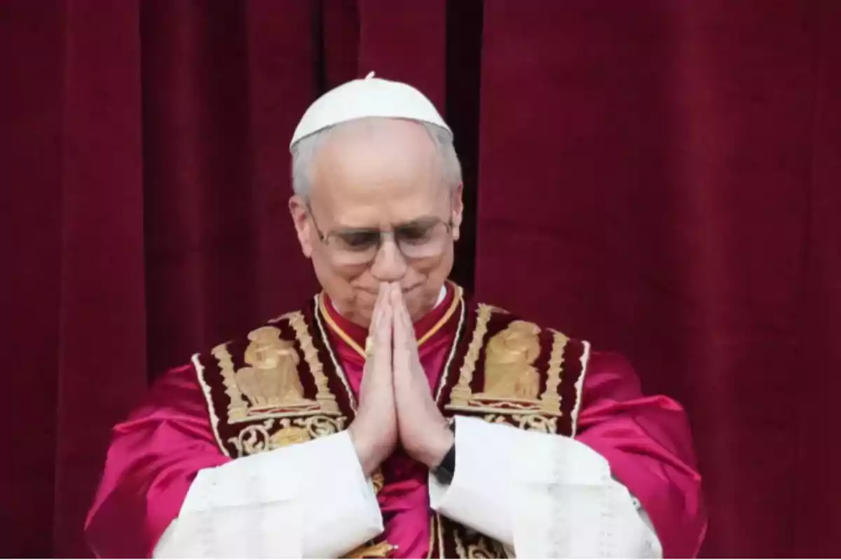 Un hombre con vestimenta religiosa y un solideo blanco junta las manos en señal de oración frente a un fondo rojo. Un hombre con vestimenta religiosa y un solideo blanco junta las manos en señal de oración frente a un fondo rojo.