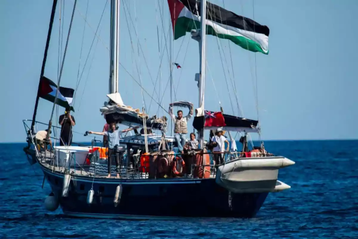 A sailboat sails on the sea with several people on board and Palestinian flags waving. A sailboat sails on the sea with several people on board and Palestinian flags waving.