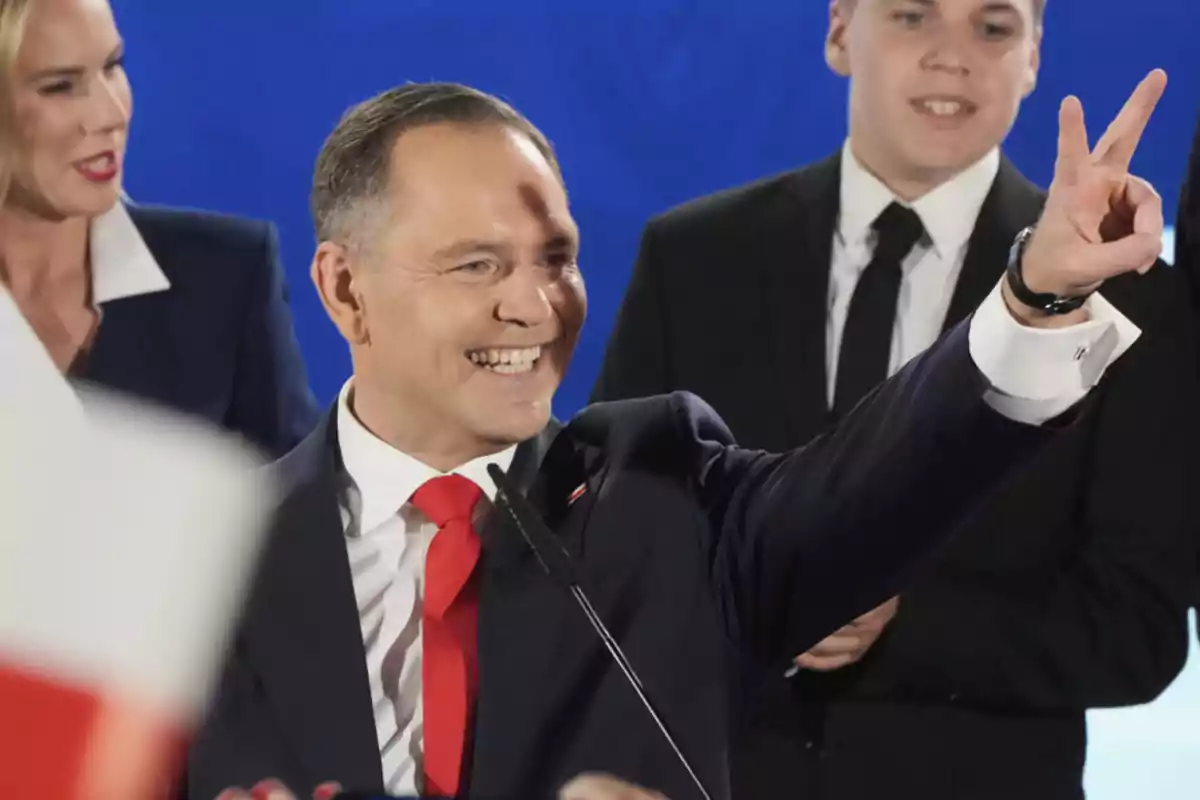 Hombre sonriente con traje y corbata roja levantando la mano y haciendo el signo de la victoria mientras habla frente a un micrófono acompañado de otras personas en un evento Hombre sonriente con traje y corbata roja levantando la mano y haciendo el signo de la victoria mientras habla frente a un micrófono acompañado de otras personas en un evento