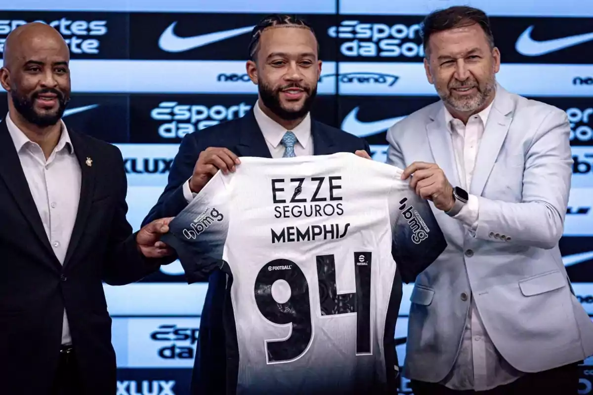 Three men in suits smile while holding a black and white soccer jersey with the name Memphis and the number 94 at a presentation event in front of a backdrop with sponsor logos. Three men in suits smile while holding a black and white soccer jersey with the name Memphis and the number 94 at a presentation event in front of a backdrop with sponsor logos.