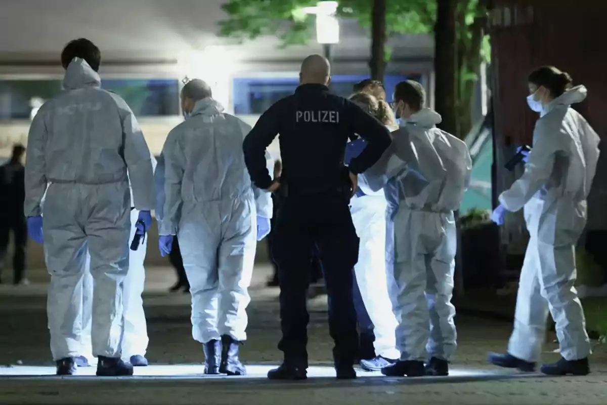 Un grupo de personas con trajes protectores y un oficial de policía se encuentran en una escena iluminada por la noche.