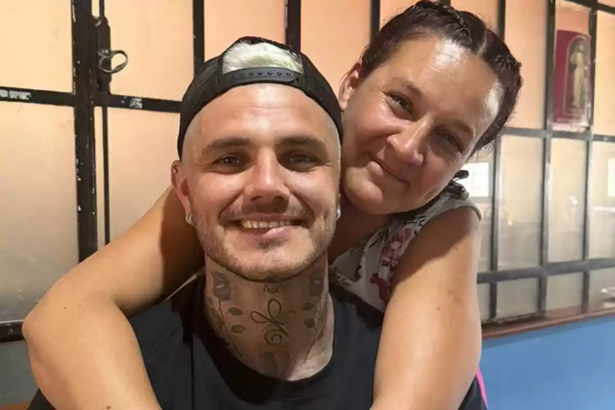 Two people smile while hugging in an indoor setting with windows in the background.