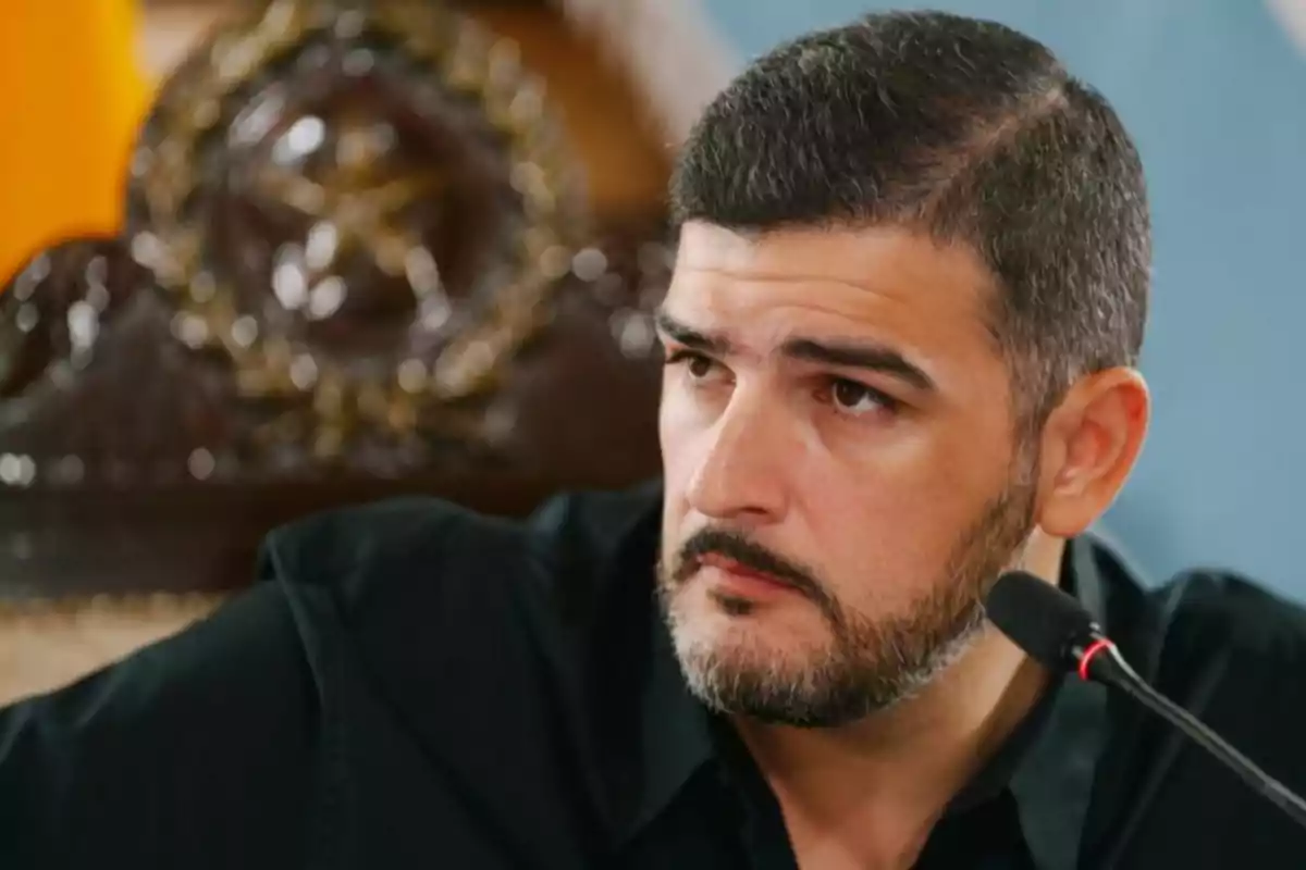 Un hombre con barba y cabello corto, vestido de negro, está hablando frente a un micrófono con un fondo decorativo. Un hombre con barba y cabello corto, vestido de negro, está hablando frente a un micrófono con un fondo decorativo.