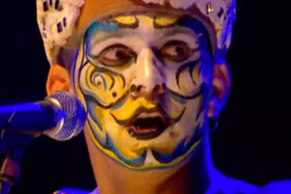 Person with colorful and detailed makeup on their face, wearing a decorated hat, in front of a microphone.