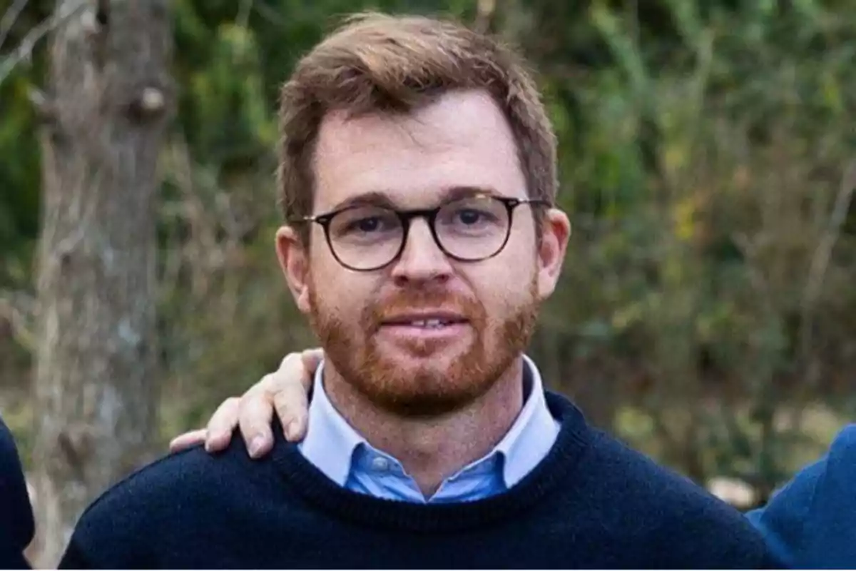 Un hombre con barba y gafas, vestido con un suéter azul oscuro y una camisa azul claro, está de pie al aire libre con un fondo de árboles y vegetación.