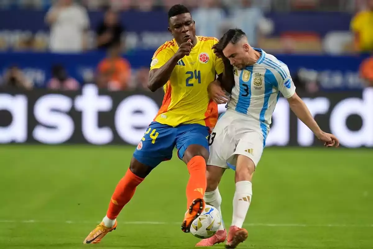 Dos jugadores de fútbol, uno con uniforme amarillo y azul y otro con uniforme blanco y celeste, compiten por el balón en un partido.