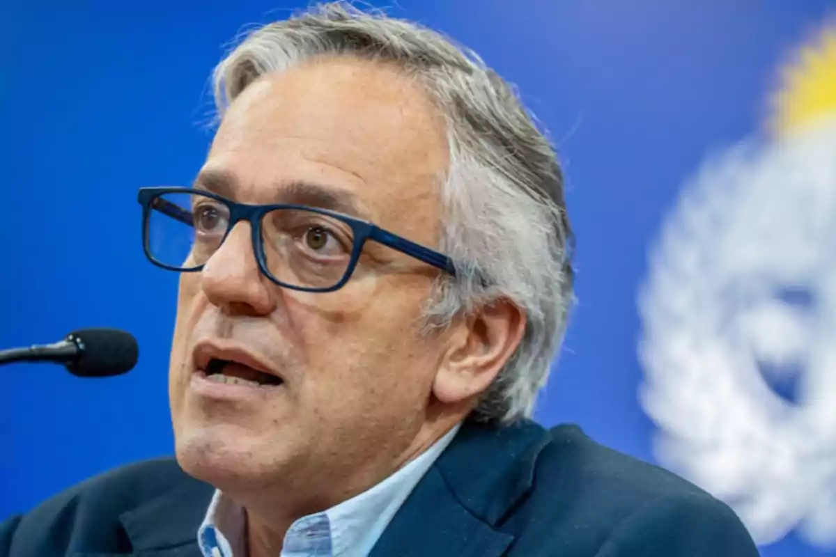 A man with glasses and gray hair speaking in front of a microphone with a blue background. A man with glasses and gray hair speaking in front of a microphone with a blue background.