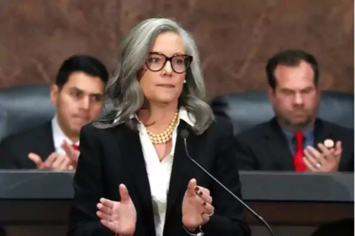 Gray-haired woman with glasses speaking into a microphone, with two men applauding in the background in a formal setting