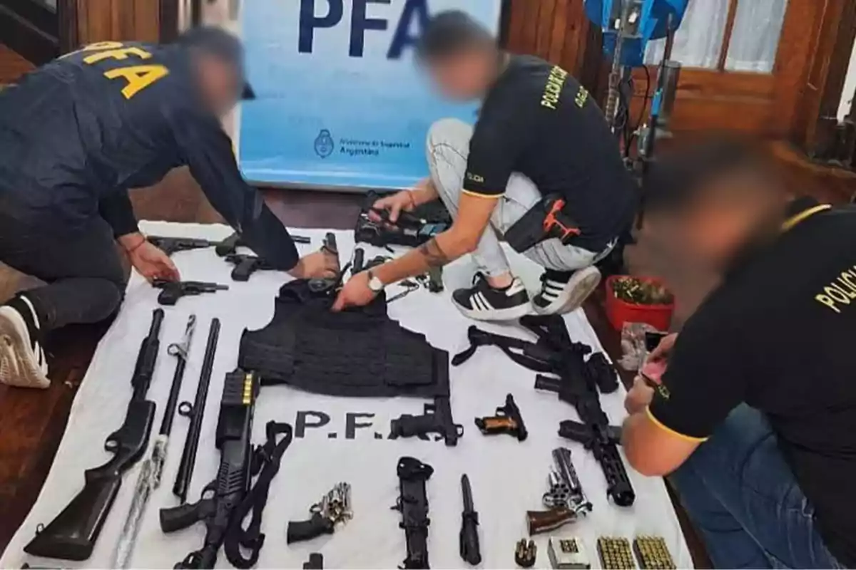 Tres personas con uniformes de la policía federal argentina revisan un arsenal de armas de fuego, municiones y un chaleco antibalas sobre una manta blanca Tres personas con uniformes de la policía federal argentina revisan un arsenal de armas de fuego, municiones y un chaleco antibalas sobre una manta blanca