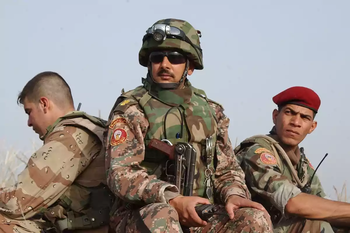 Three soldiers in camouflage uniforms and military gear are sitting outdoors.