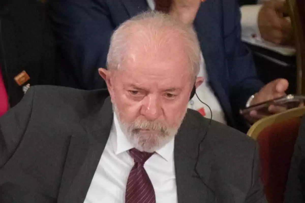 Older man with gray hair and beard wears a formal suit and tie while listening alertly with an earpiece at an event.