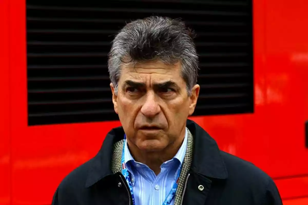 Hombre de cabello canoso con expresión seria frente a un fondo rojo. Hombre de cabello canoso con expresión seria frente a un fondo rojo.