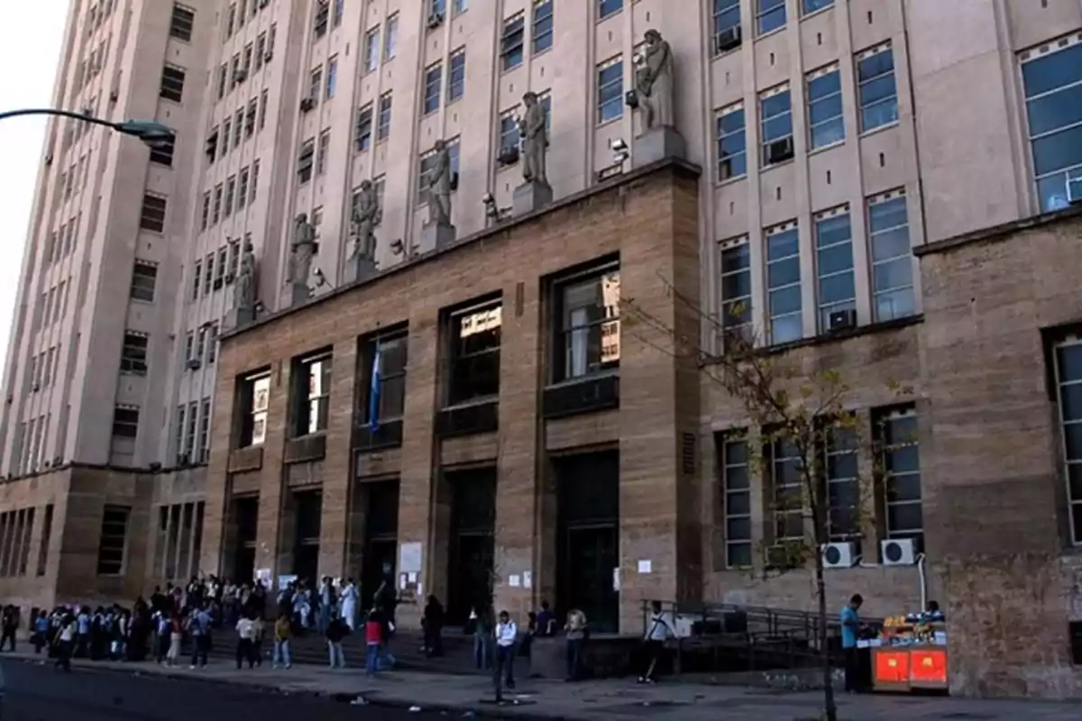 Fachada de un edificio grande con esculturas en la parte superior y personas reunidas en la entrada.