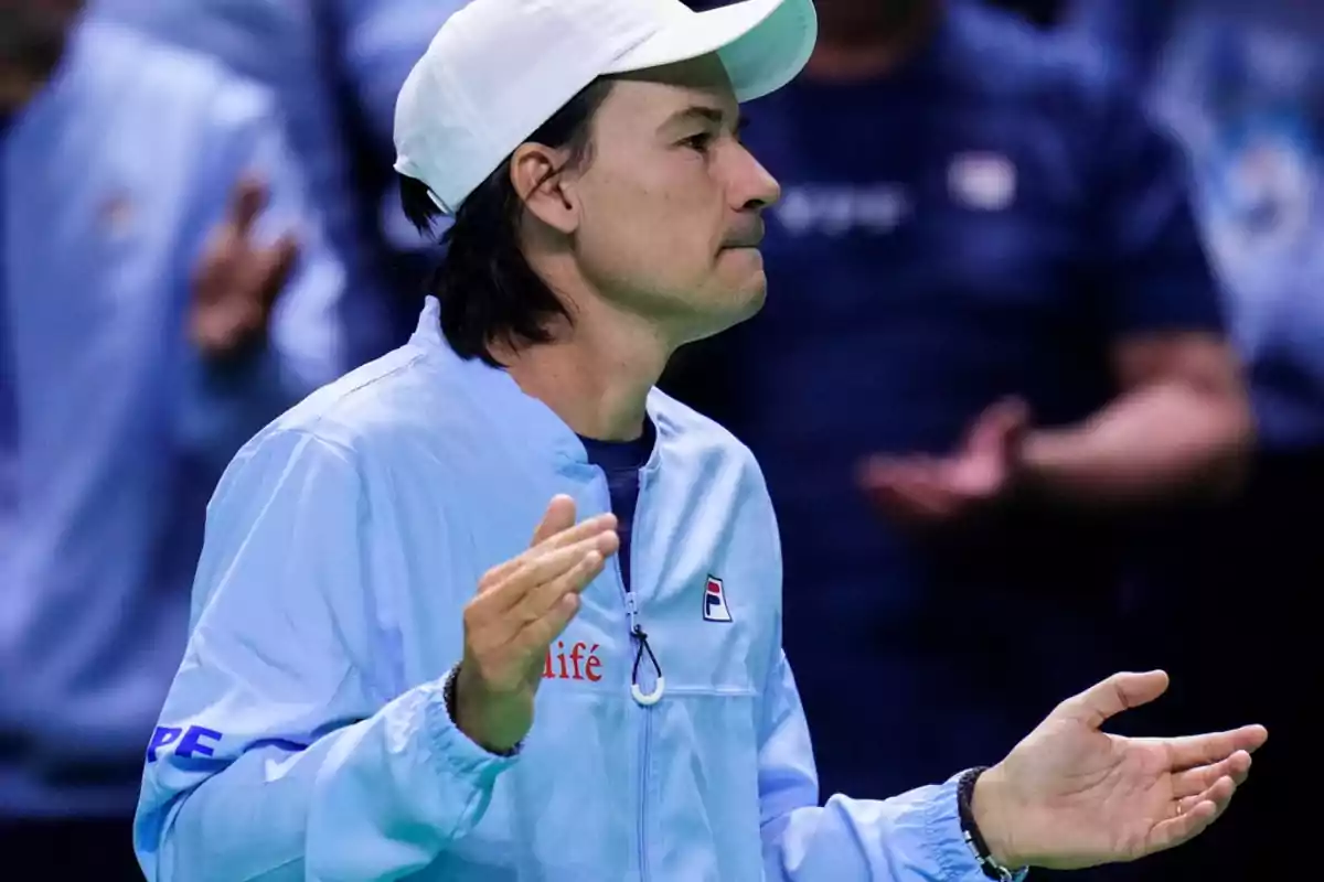 Un hombre con gorra blanca y chaqueta azul aplaude en un evento deportivo.