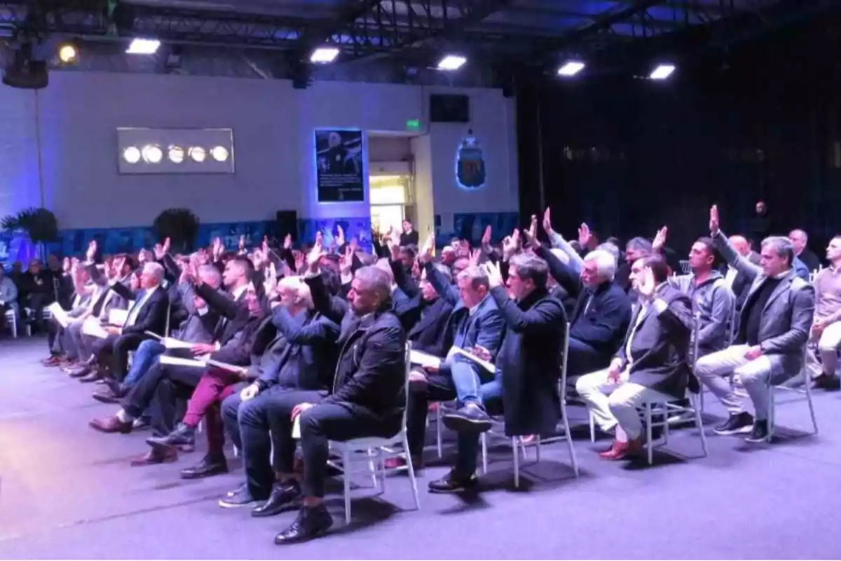 A group of people sitting in chairs raising their hands at an event in a well-lit room. A group of people sitting in chairs raising their hands at an event in a well-lit room.