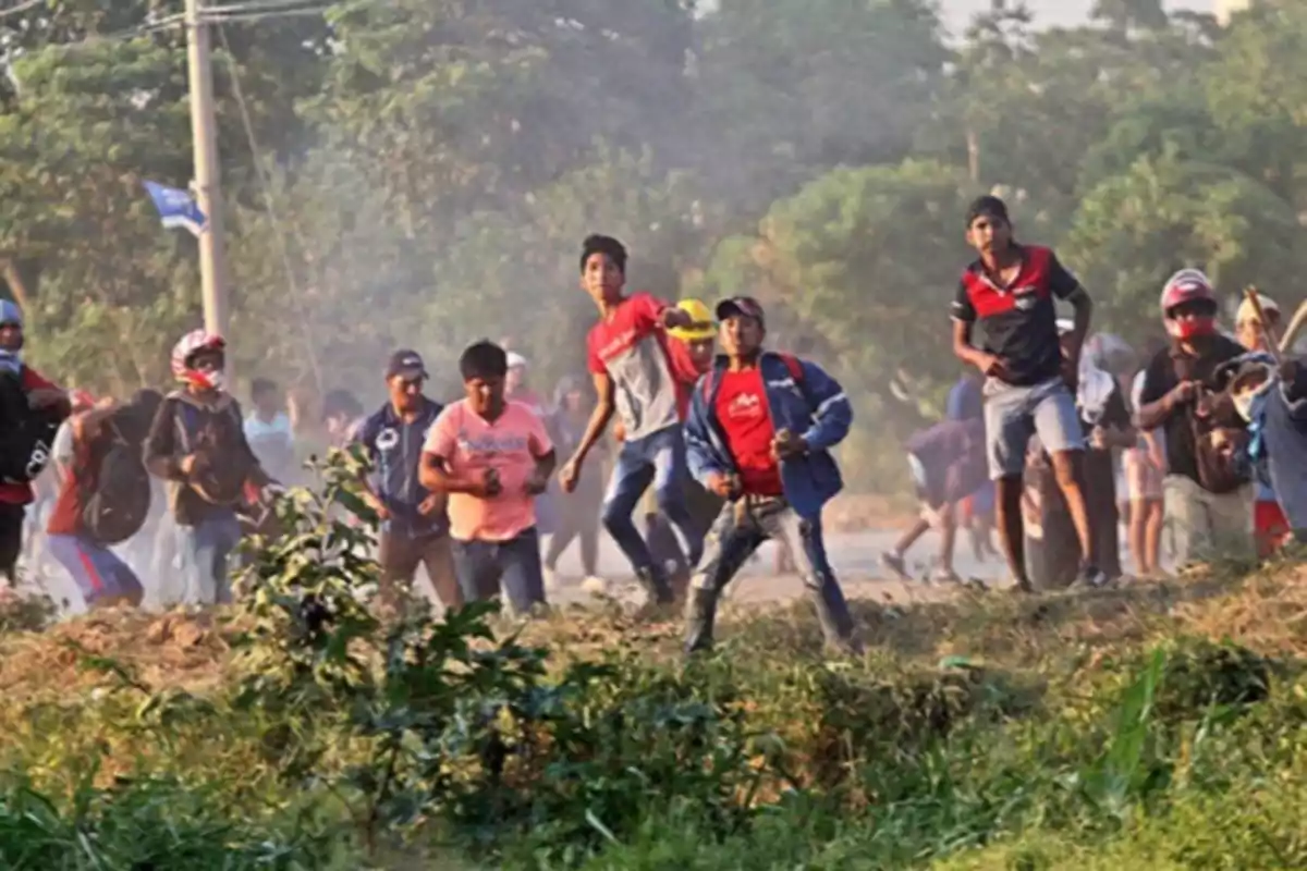 Un grupo de personas corriendo en un entorno al aire libre con vegetación y humo en el fondo.