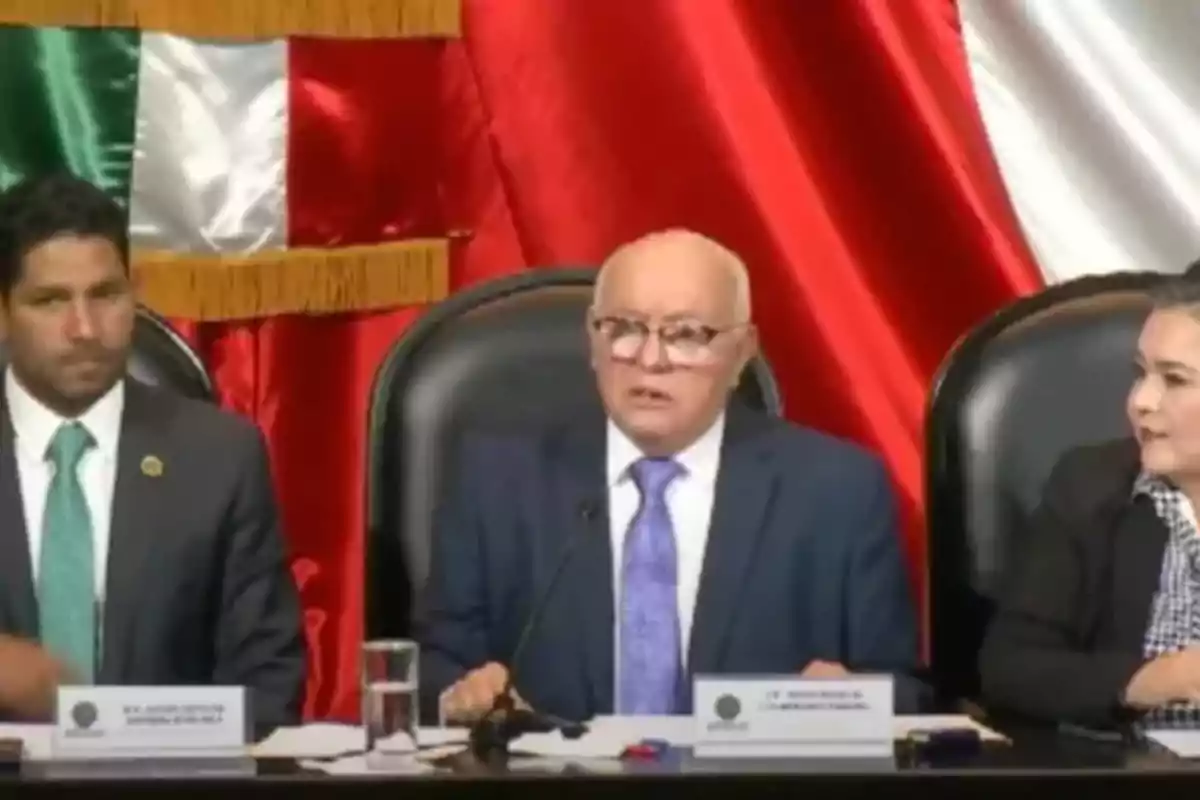 Tres personas sentadas en una mesa con micrófonos frente a una bandera de México.