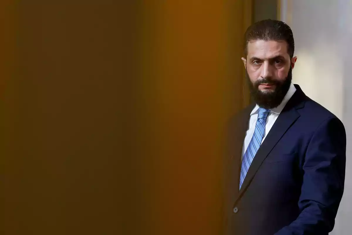 Hombre con traje oscuro y corbata azul de pie frente a un fondo marrón. Hombre con traje oscuro y corbata azul de pie frente a un fondo marrón.