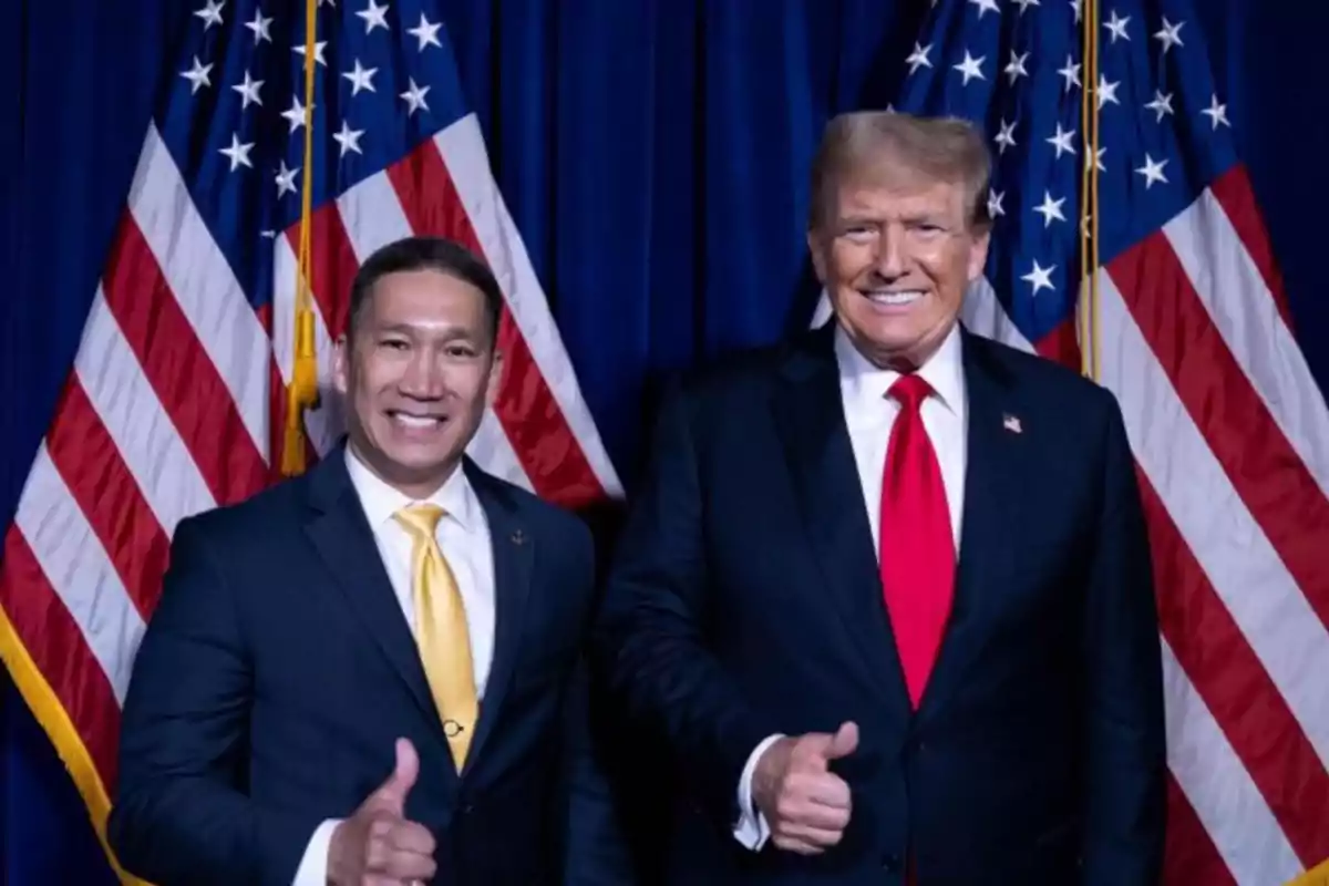 Two people smiling and giving a thumbs up in front of United States flags.