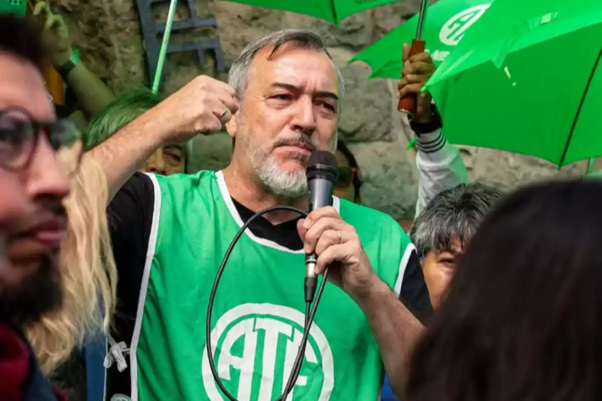 Un hombre con chaleco verde y micrófono habla en una manifestación rodeado de personas con paraguas verdes.