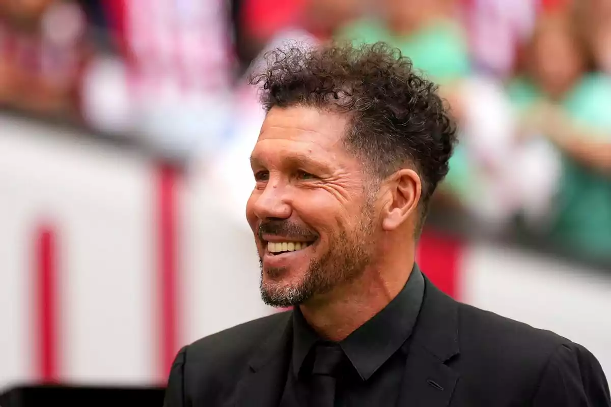 Un hombre sonriente con cabello rizado y barba viste un traje negro en un entorno deportivo. Un hombre sonriente con cabello rizado y barba viste un traje negro en un entorno deportivo.