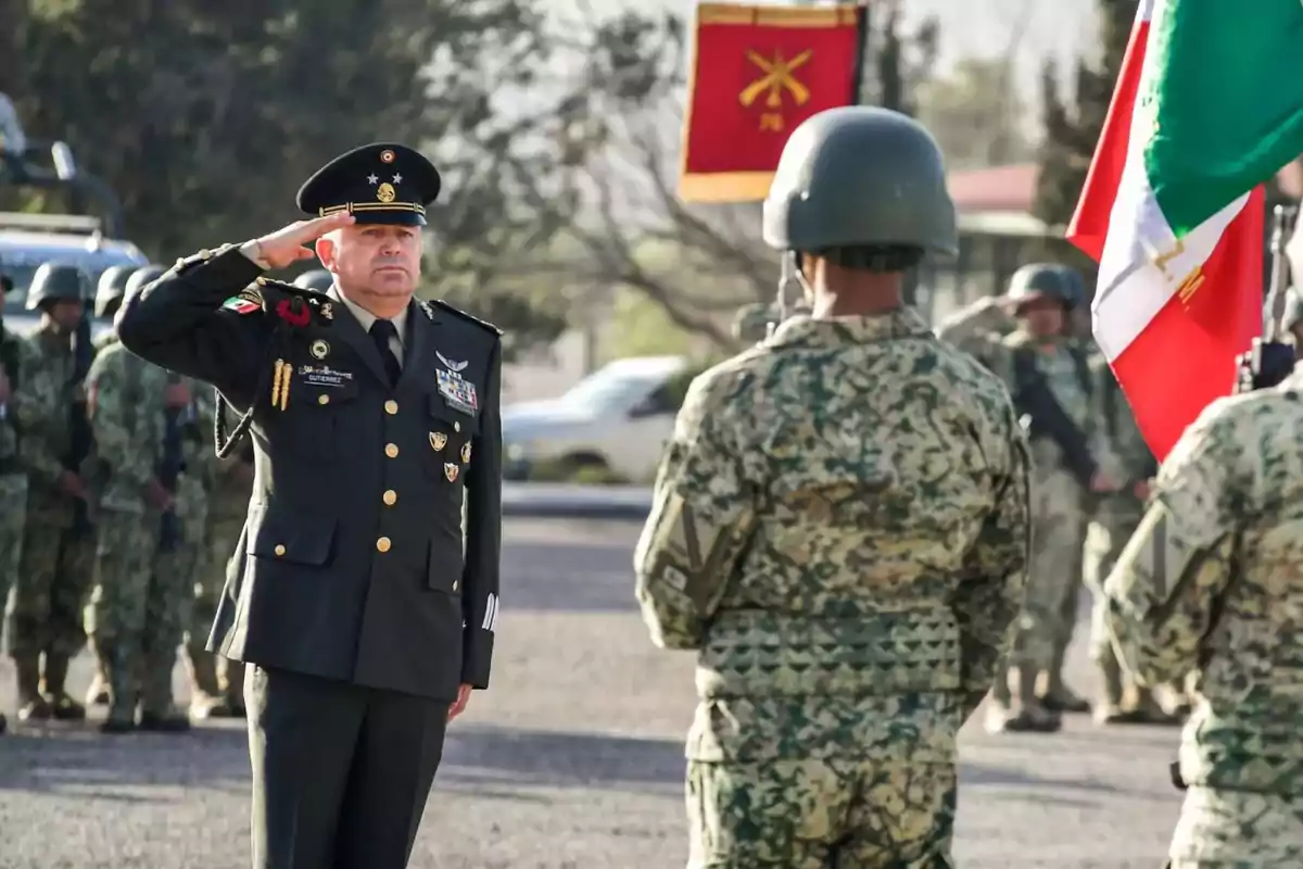 Un militar de alto rango saluda a un grupo de soldados en formación, con banderas y uniformes de camuflaje en un entorno al aire libre.