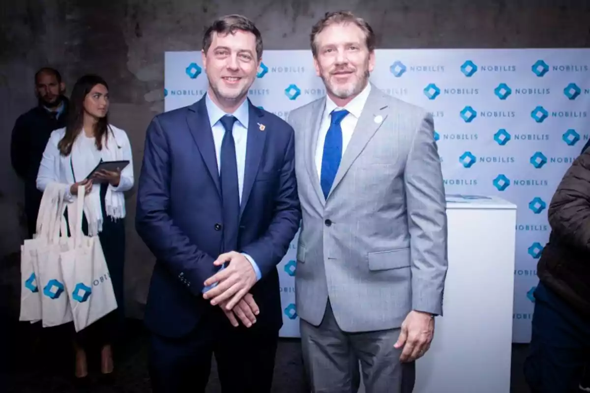 Dos hombres vestidos con traje posan sonrientes en un evento corporativo con fondo de logotipos de Nobilis y personas en segundo plano Dos hombres vestidos con traje posan sonrientes en un evento corporativo con fondo de logotipos de Nobilis y personas en segundo plano