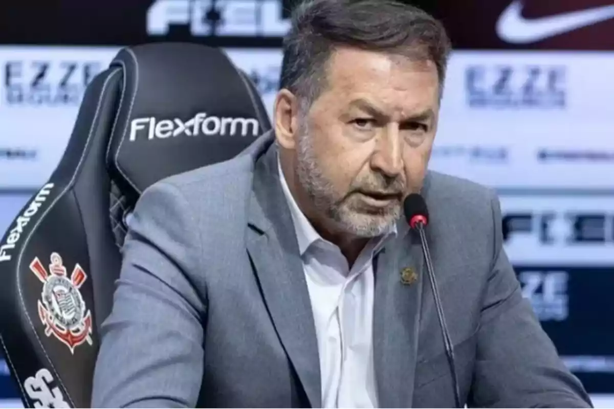 Man in a gray suit speaking at a press conference seated in a chair with the Corinthians club logo Man in a gray suit speaking at a press conference seated in a chair with the Corinthians club logo