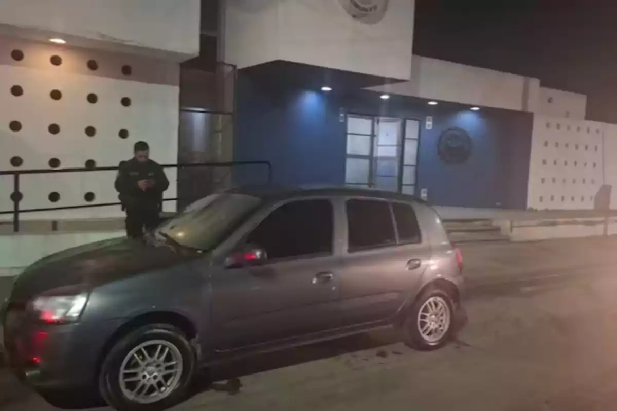 A gray car parked in front of a modern building with white walls and circular details, while a person in a dark uniform stands near the vehicle using a mobile phone.