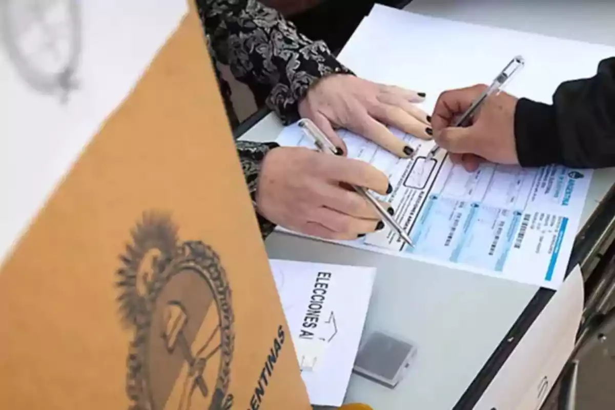 People filling out forms at a voting table with a cardboard ballot box in front.