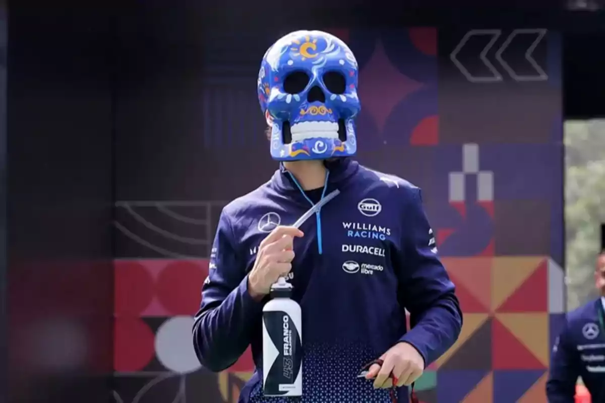 A person in a Williams Racing uniform wears a decorated skull mask and holds a water bottle against a colorful background. A person in a Williams Racing uniform wears a decorated skull mask and holds a water bottle against a colorful background.
