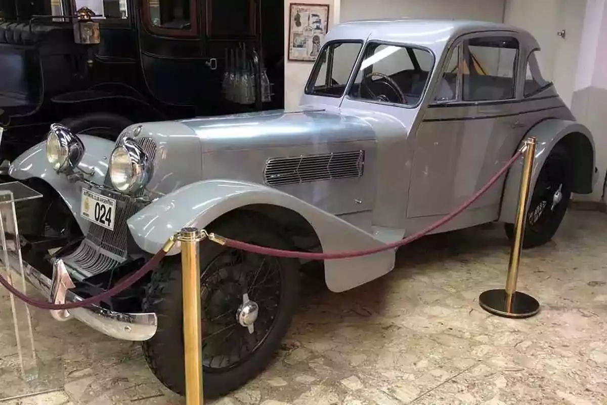 Un automóvil clásico de color plateado exhibido en un museo, rodeado por una cuerda de seguridad. Un automóvil clásico de color plateado exhibido en un museo, rodeado por una cuerda de seguridad.