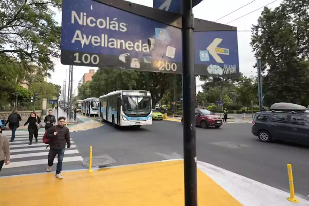 Personas cruzan una calle en una intersección urbana con señalización vial y varios autobuses y autos circulando, rodeados de árboles y edificios. Personas cruzan una calle en una intersección urbana con señalización vial y varios autobuses y autos circulando, rodeados de árboles y edificios.