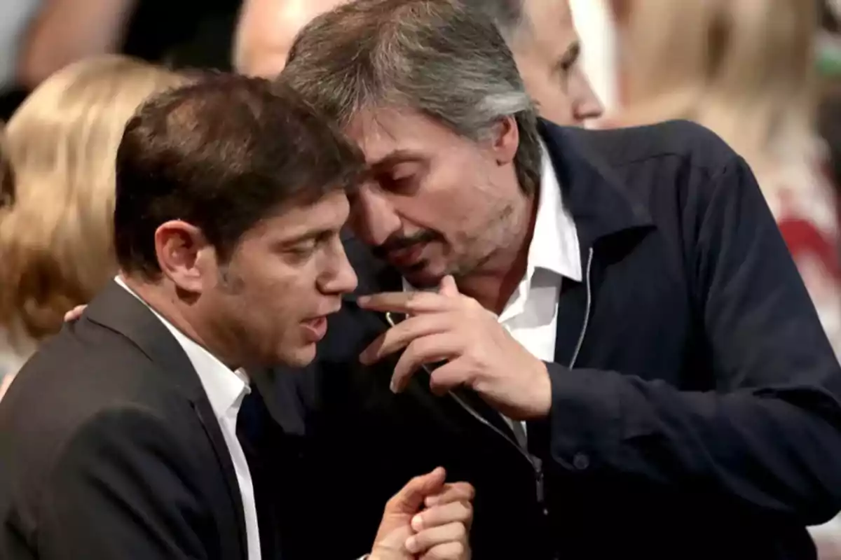 Dos hombres conversan de cerca en un evento, uno de ellos susurra al oído del otro mientras gesticula con la mano.