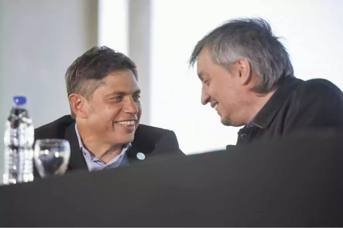 Dos personas conversando y sonriendo en un evento con una botella de agua y un vaso en la mesa.