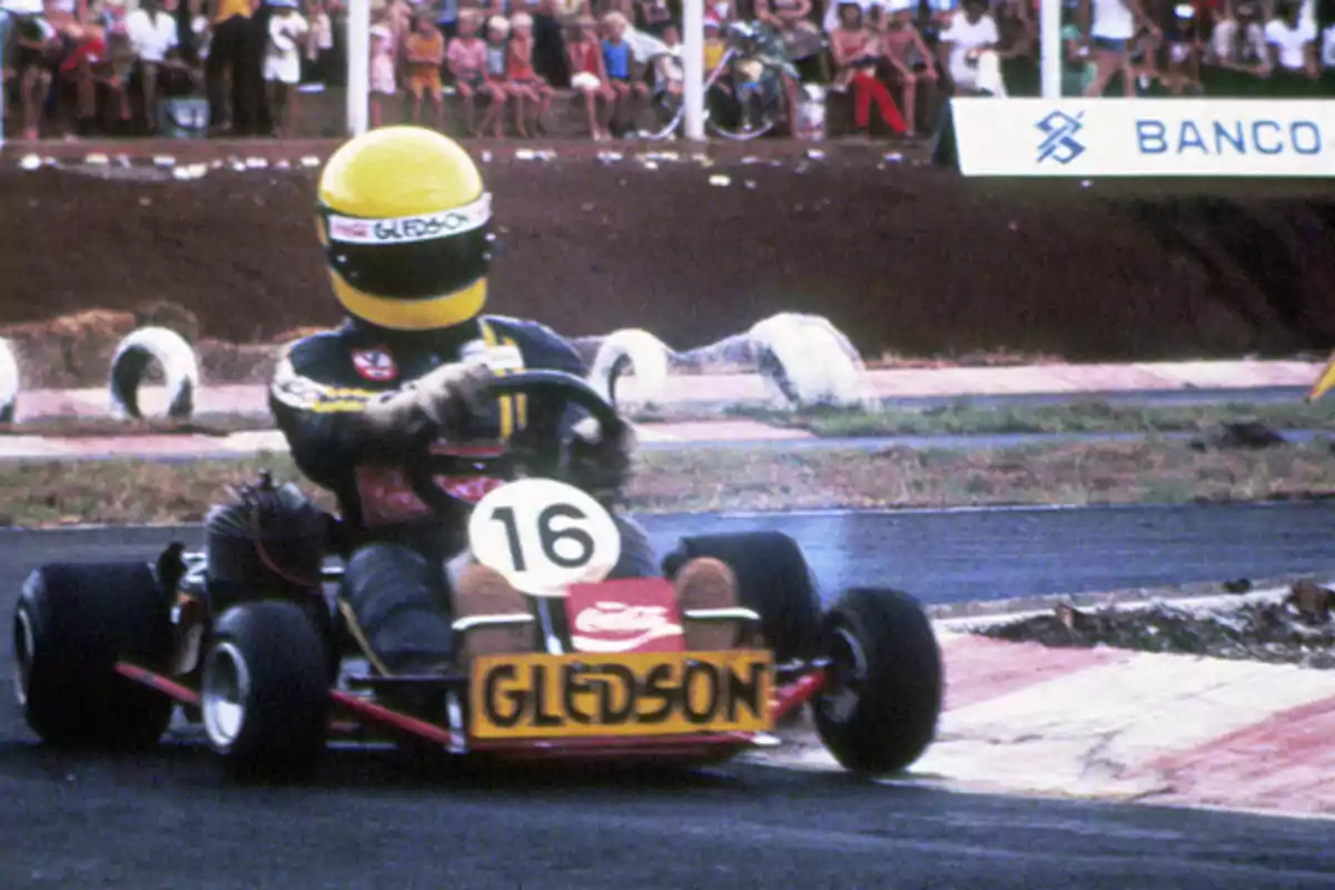 A karting driver with a yellow helmet competes on a track while the audience watches from the stands. A karting driver with a yellow helmet competes on a track while the audience watches from the stands.