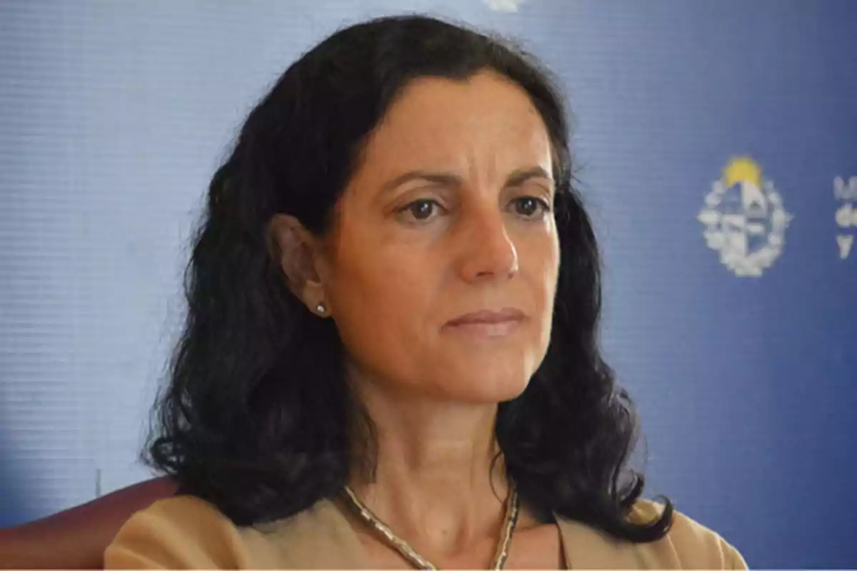 Dark-haired, wavy-haired woman with a serious expression in front of a blue background with a blurred institutional emblem.