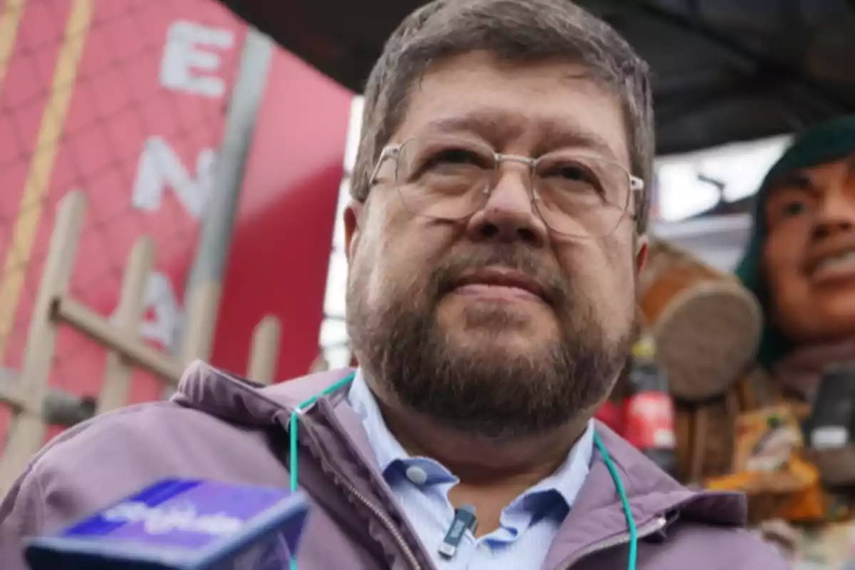 A man with glasses and a beard is outdoors, with a blue microphone in the foreground and a blurred background that includes a colorful figure. A man with glasses and a beard is outdoors, with a blue microphone in the foreground and a blurred background that includes a colorful figure.