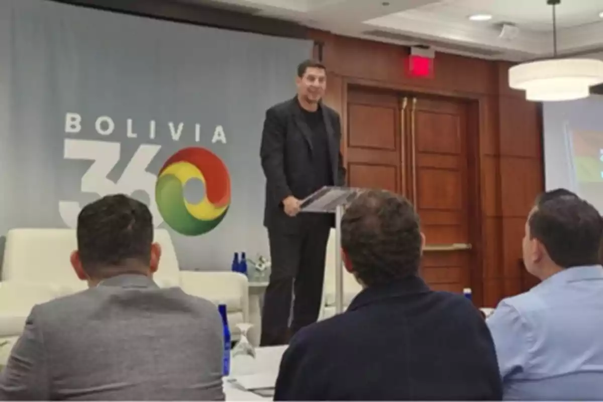 Un hombre de pie en un podio habla frente a un grupo de personas sentadas en una sala de conferencias con un cartel que dice "Bolivia 360" en el fondo.