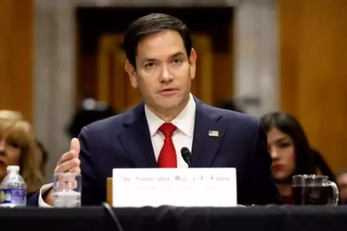 A man in a dark suit and red tie speaks into a microphone during a hearing in a formal setting. A man in a dark suit and red tie speaks into a microphone during a hearing in a formal setting.