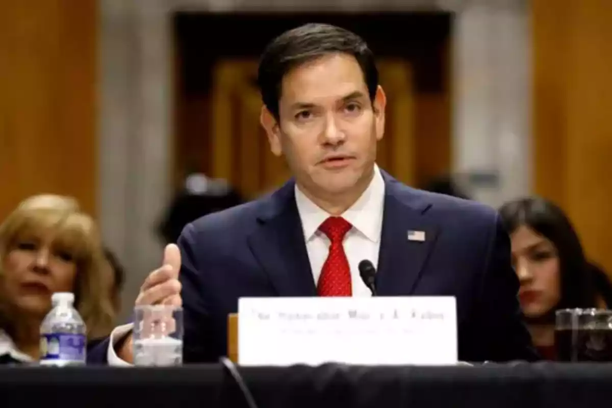 Dark-haired man in a blue suit and red tie speaking into a microphone during an official hearing, with people seated in the background. Dark-haired man in a blue suit and red tie speaking into a microphone during an official hearing, with people seated in the background.