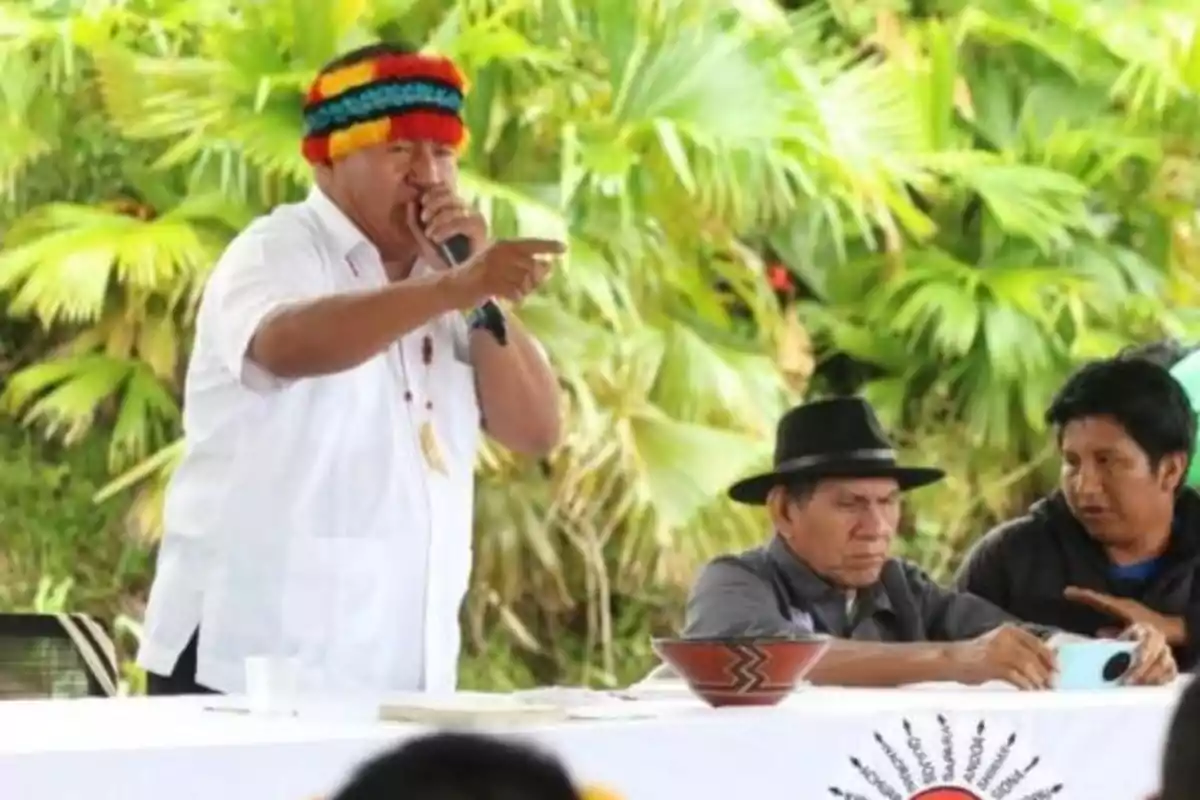 A man with a colorful crown speaks into a microphone while pointing, joined by two people sitting next to him, in an outdoor setting with vegetation in the background.