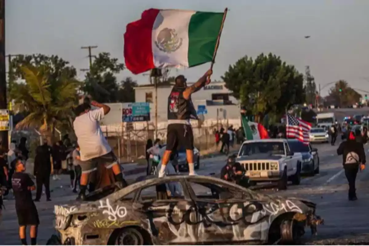 Hombre de pie sobre un auto quemado y grafiteado, ondeando una bandera de México en medio de una calle con más personas y vehículos al fondo Hombre de pie sobre un auto quemado y grafiteado, ondeando una bandera de México en medio de una calle con más personas y vehículos al fondo