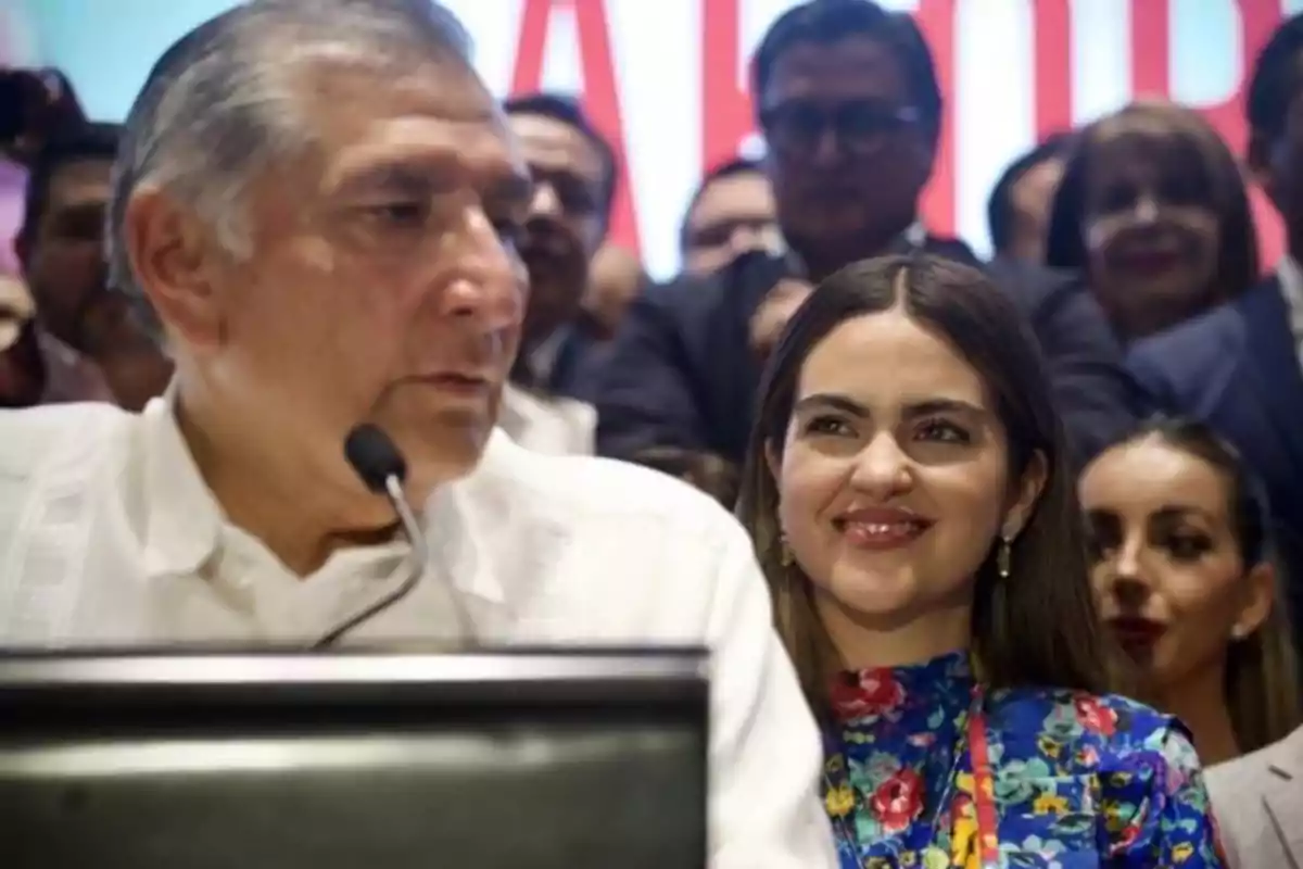 A man speaking into a microphone while a woman smiles beside him at an event with several people in the background. A man speaking into a microphone while a woman smiles beside him at an event with several people in the background.