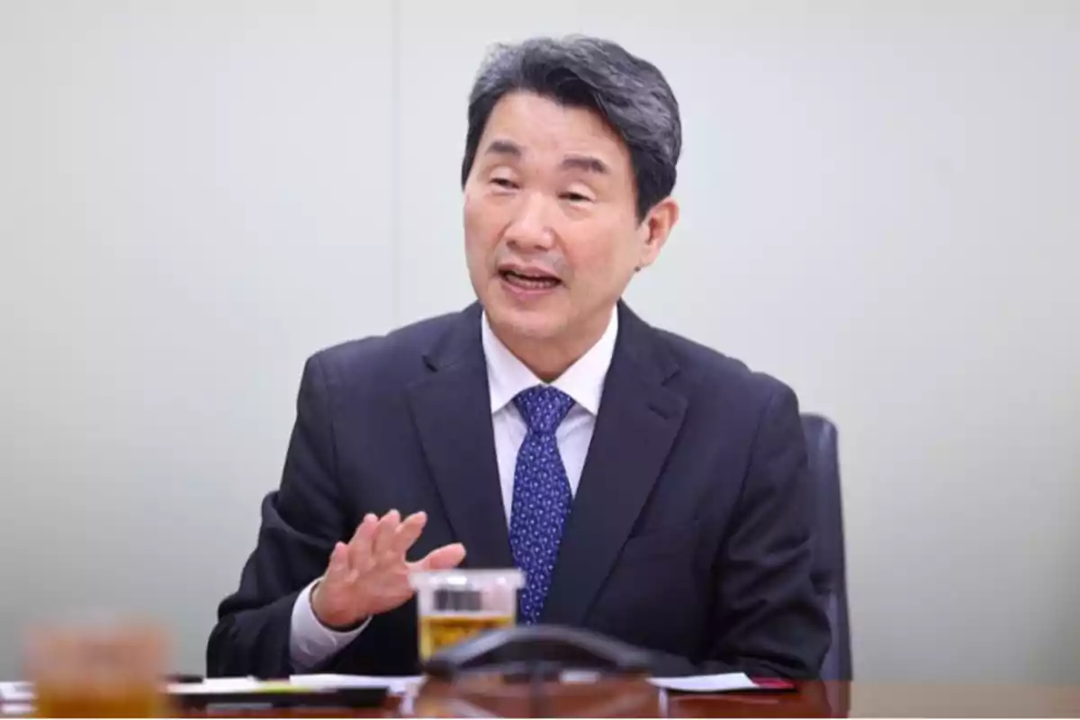 A man in a dark suit and blue tie speaks gesturing with his hand in a meeting room. A man in a dark suit and blue tie speaks gesturing with his hand in a meeting room.