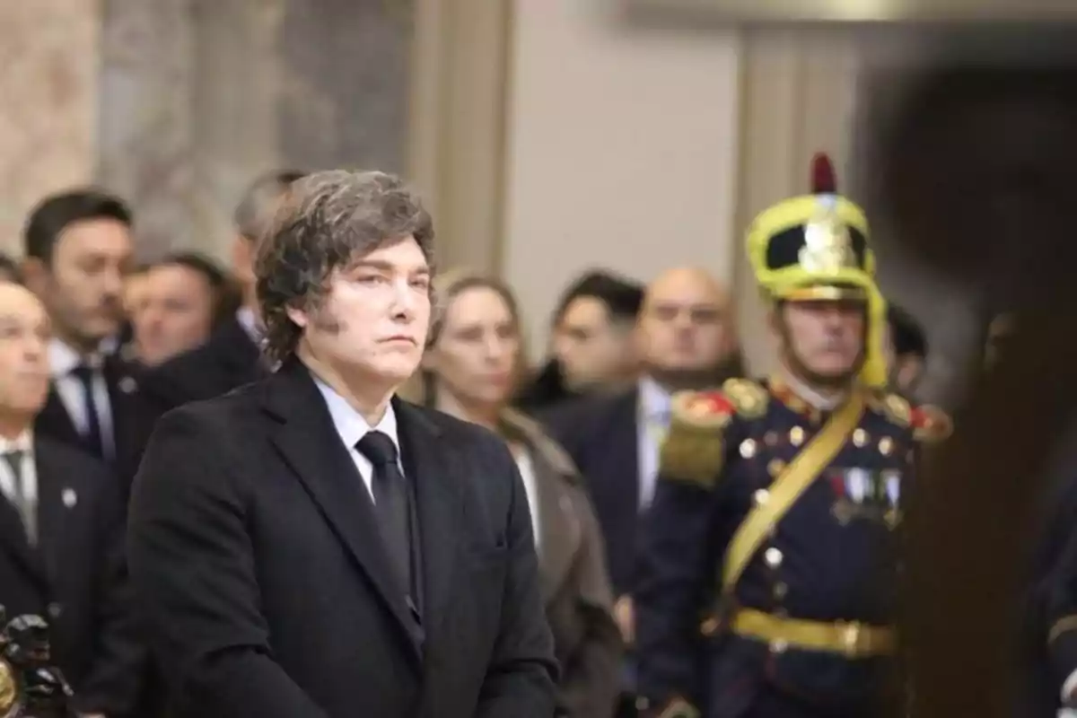 Wavy-haired man in a dark suit at a formal event with uniformed people and attendees in the background Wavy-haired man in a dark suit at a formal event with uniformed people and attendees in the background